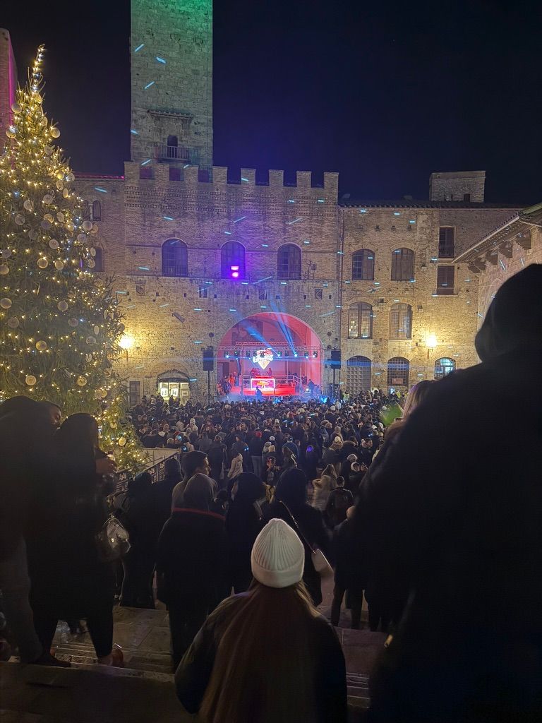 Folla di spalle verso il palco di Capodanno a San Gimignano, albero e torre illuminati sulla piazza