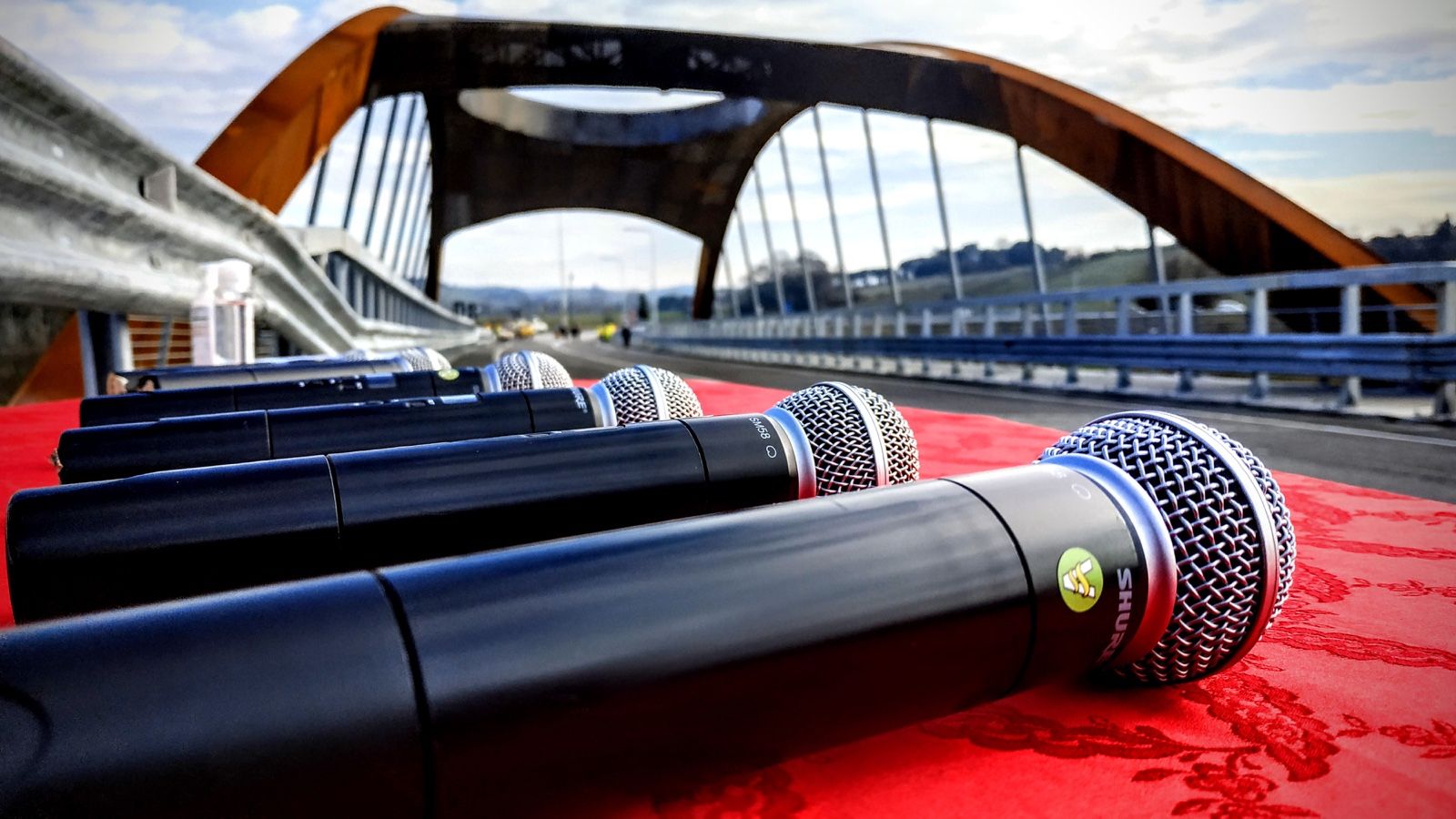 Fila di radiomicrofoni pronti sul tavolo rosso di un'inaugurazione, ponte sullo sfondo