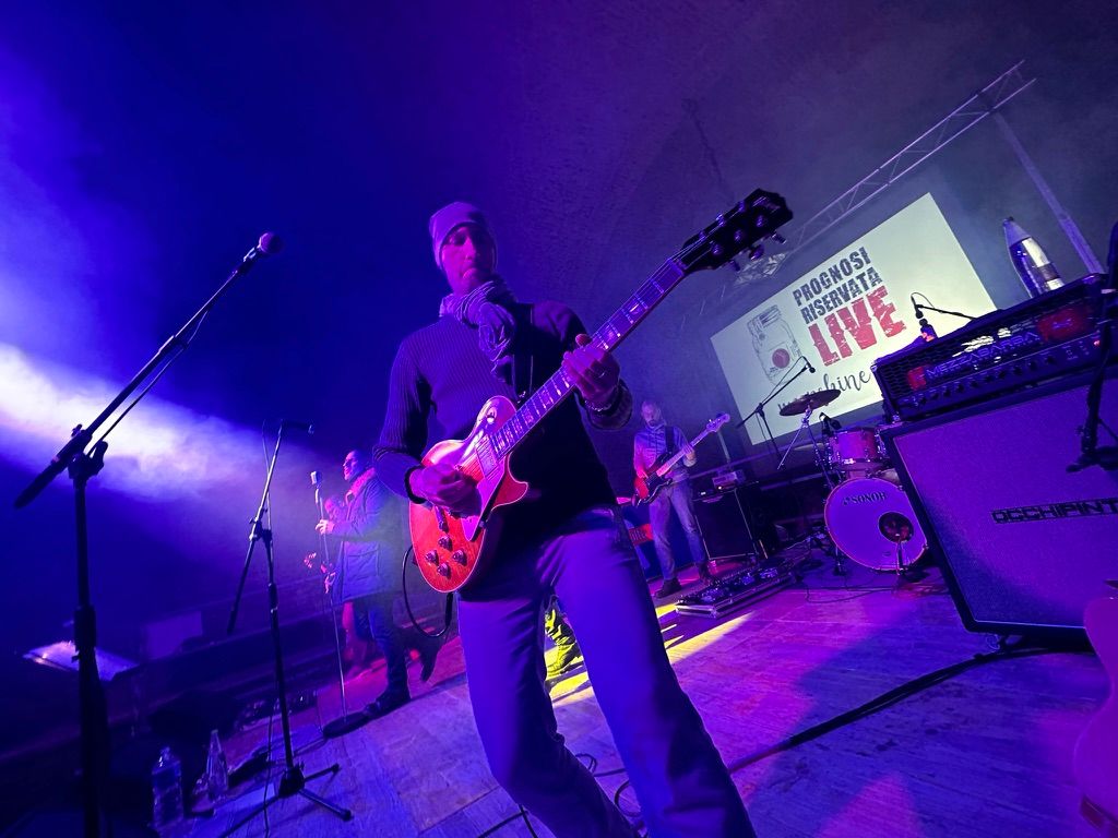 Chitarrista in primo piano dal basso su palco viola, band e truss sullo sfondo
