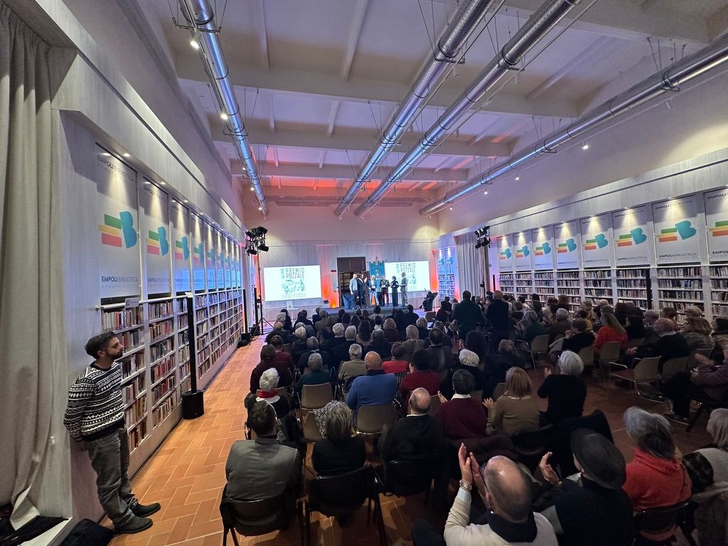 Sala della biblioteca gremita vista dal fondo, palco e schermi in fondo alla prospettiva