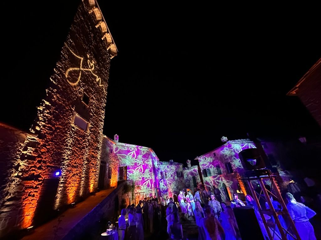 Borgo toscano di notte con monogramma e proiezioni floreali sulle facciate, festa in corso