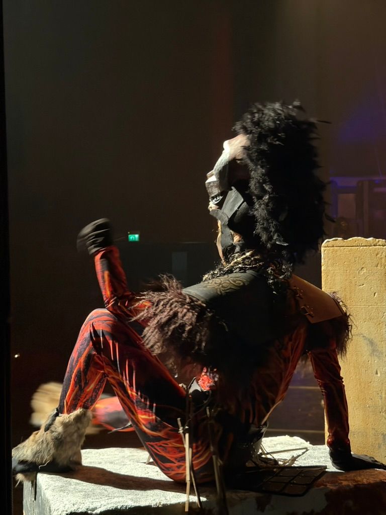 Performer in costume di scena elaborato sul blocco scenografico, braccio alzato nel controluce